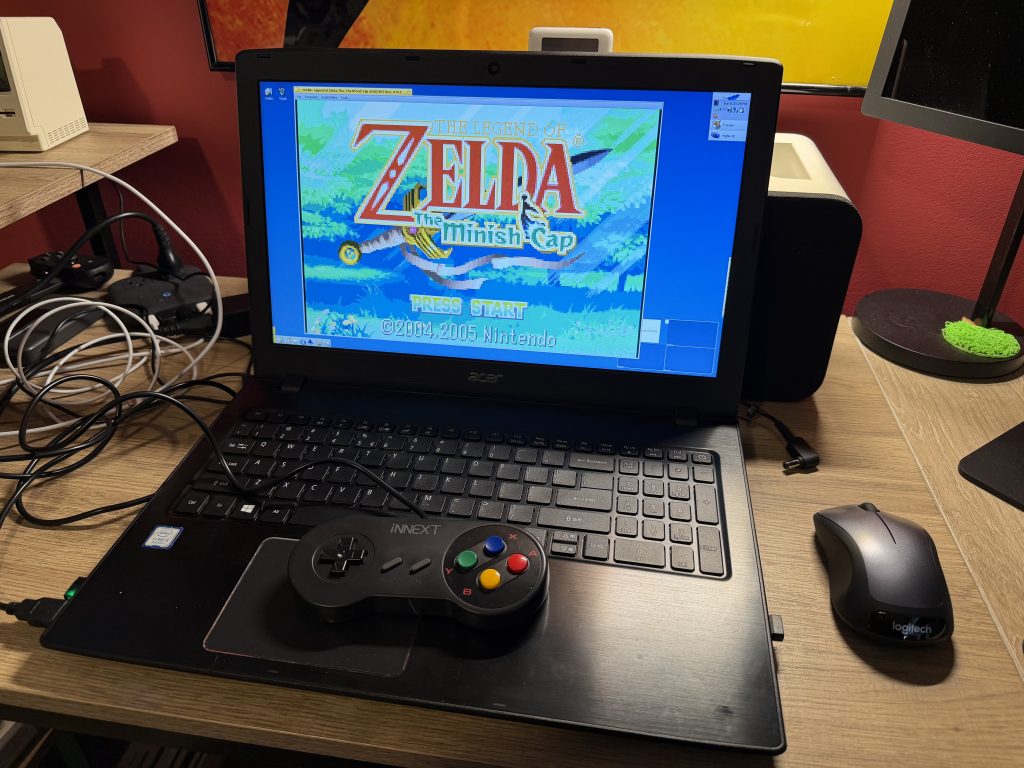 a laptop sitting on a desk. a gameboy advance emulator running zelda - the minish cap is shown on screen. a super nintendo-style controller is plugged in