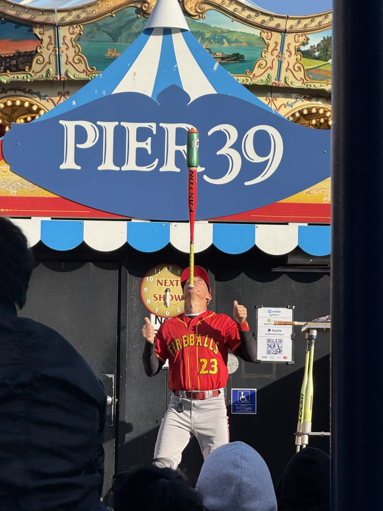 a man balances a bat on his chin in front of a sign that reads "pier 39"