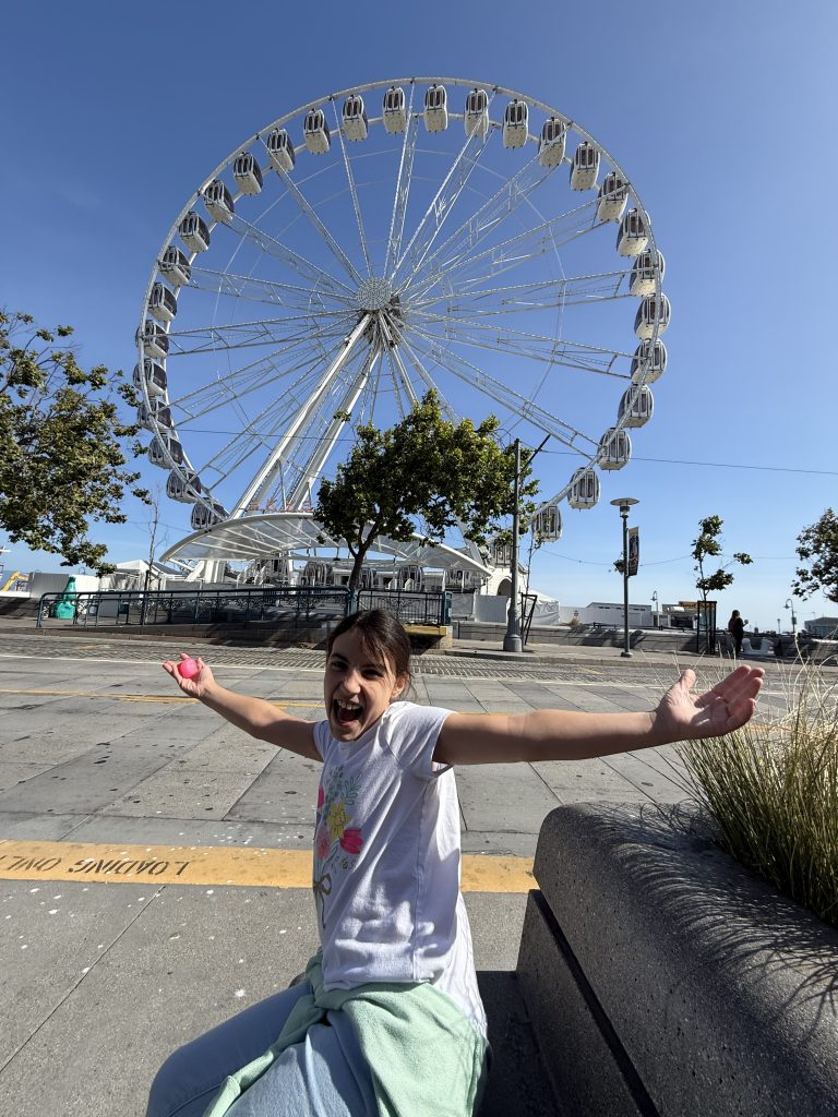 a girl sits in front of a ferris wheel with her arms spread wide