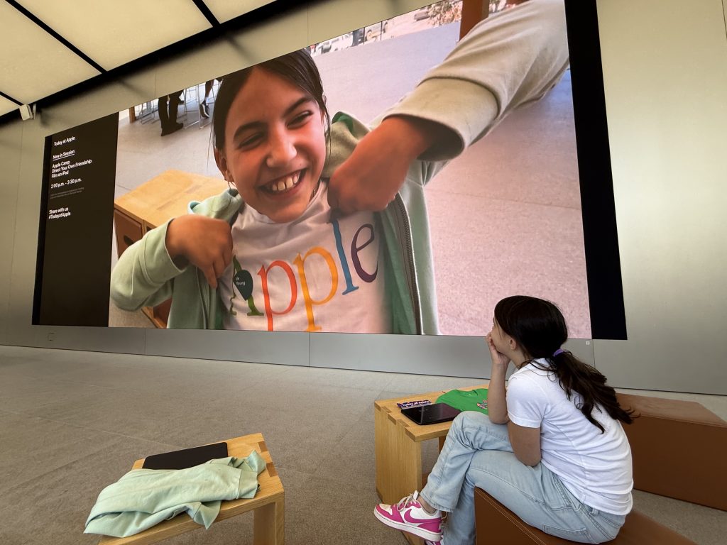 a girl sits looking at herself on a large screen inside an Apple store