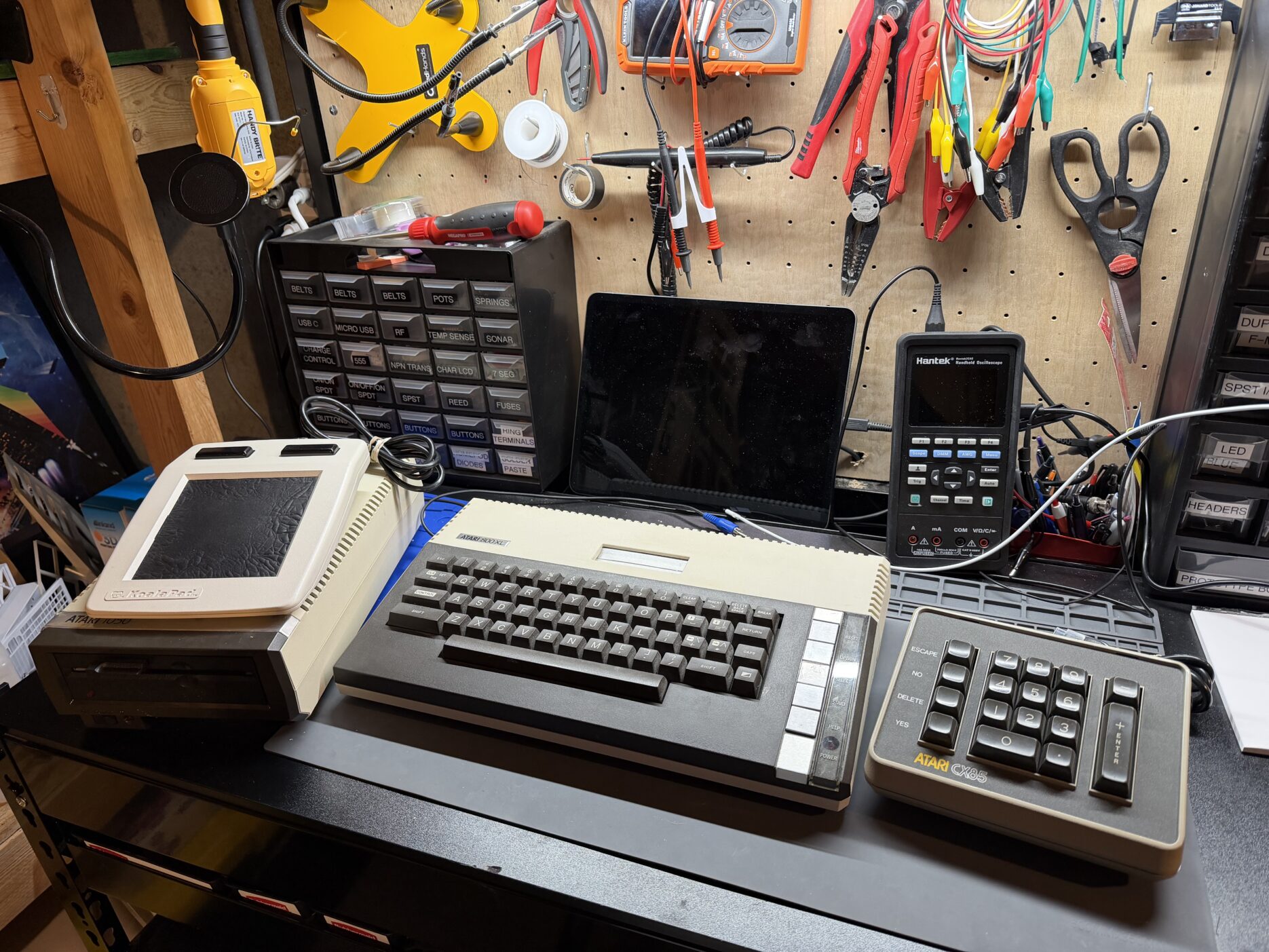an atari 800xl computer sits on a workbench with a keypad and disk drive on either side. the disk drive has a koala pad on top of it. surrounded by various electronics repair tools