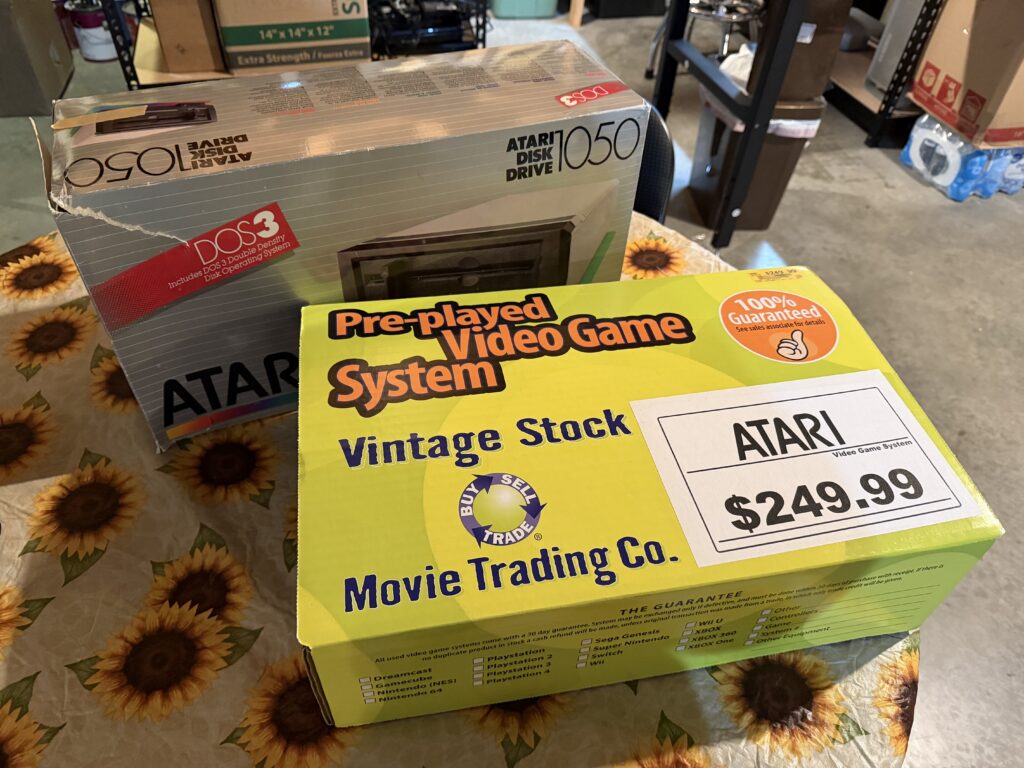 An Atari 1050 disk drive box sitting behind a green vintage stock box with a label that says "Atari $249.99"