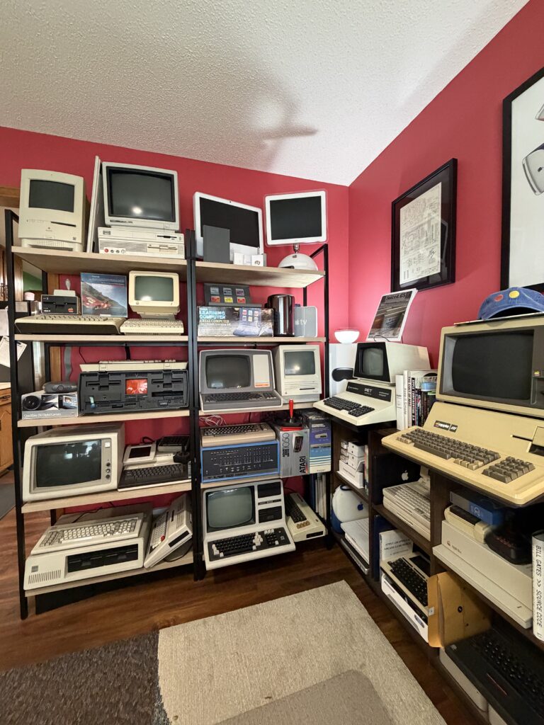 A room with bright red walls is filled with vintage computers and electronics displayed on shelves. The collection includes bulky beige desktop computers, CRT monitors, early Apple models, an Atari box, and other retro keyboards and hardware. A signed Apple I schematic and a few books are also visible
