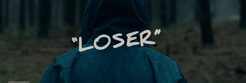 A person stands in the woods, their back to the camera. A damp raincoat covers their head and back. the word "Loser" is printed over the person
